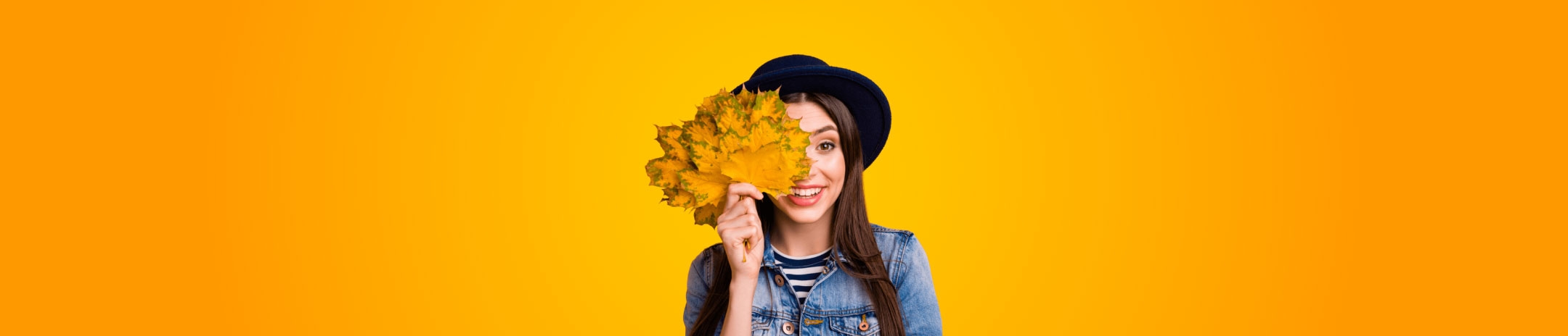 A woman in a striped shirt, black hat and jean jacket, holding up fall leaves in front of a gradient orange background.