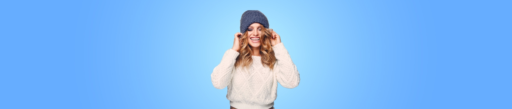 Girl smiling in cream sweater and blue knit hat, in front of a light blue gradient background